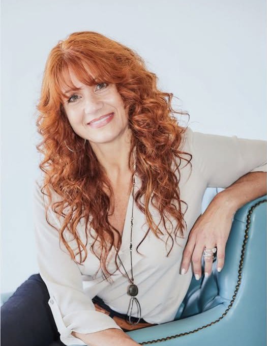 Adrianne Luban, smiling at the camera, sitting on the arm of a blue leather chair.